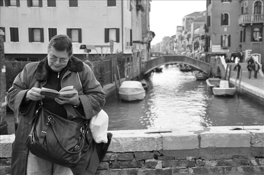 An old woman waits for the opening of a soup kitchen reading a prayer book