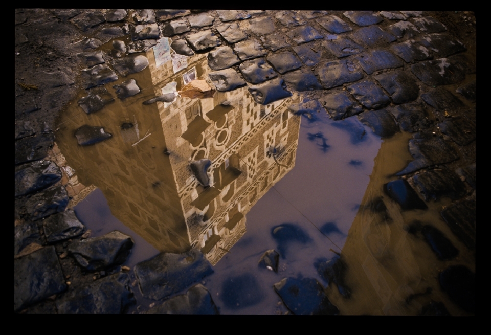 A momentary reflection of a traditional Yemeni towerhouse and it's political posters in a puddle  over the cobblestones. Political posters of President Saleh can be seen, a man whose own world would be turned upside down during the Arab Spring.