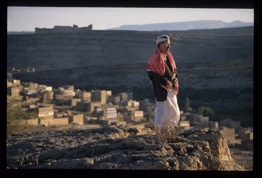 A Yemeni villager surveys his homeland, the ancient fortress above that once protected the valley and his village below, glowing in the last beams of the sun to penetrate the narrow gorge.Within a few years his long-time president will be ousted and US drone strikes will threaten this tranquil landscape.