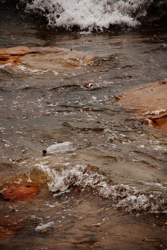 As I gaze out along the entire surface of the sea where the waves crash in on the shore, countless plastic bottles, cans and other trash create a black sludge-like body of debris that churns and toils in the chaos caused by a large storm. 