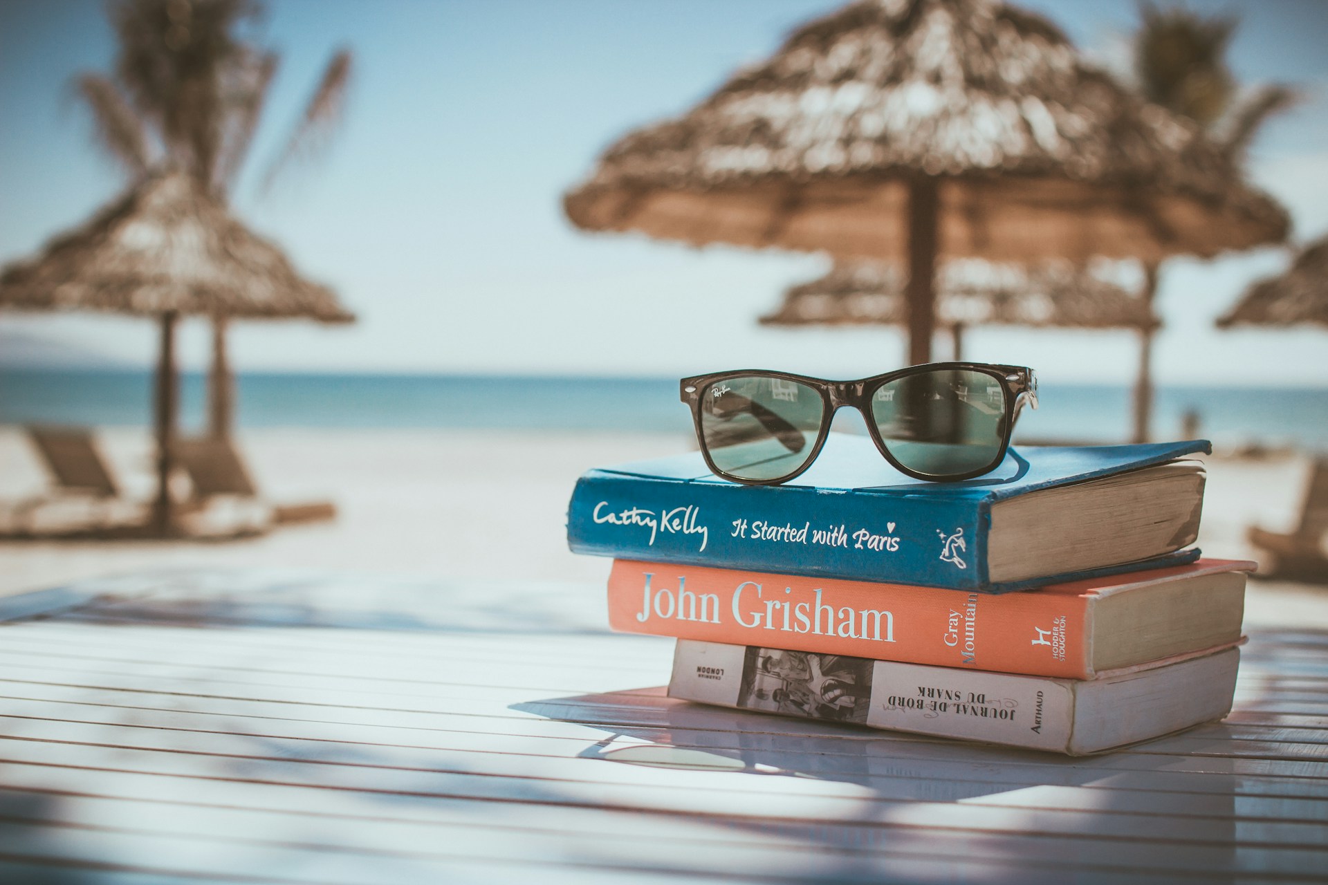 A pile of books stacked on a sunny beach, with clear blue water and straw parasols