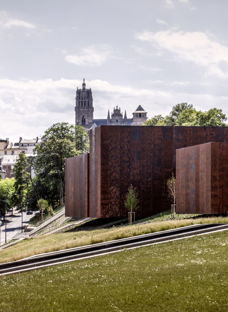 Vista lateral del museo Soulages, con una apariencia similar a la de un contenedor en un camino inclinado que lleva a un parque local, lo que crea una conexión entre el área verde y la ciudad