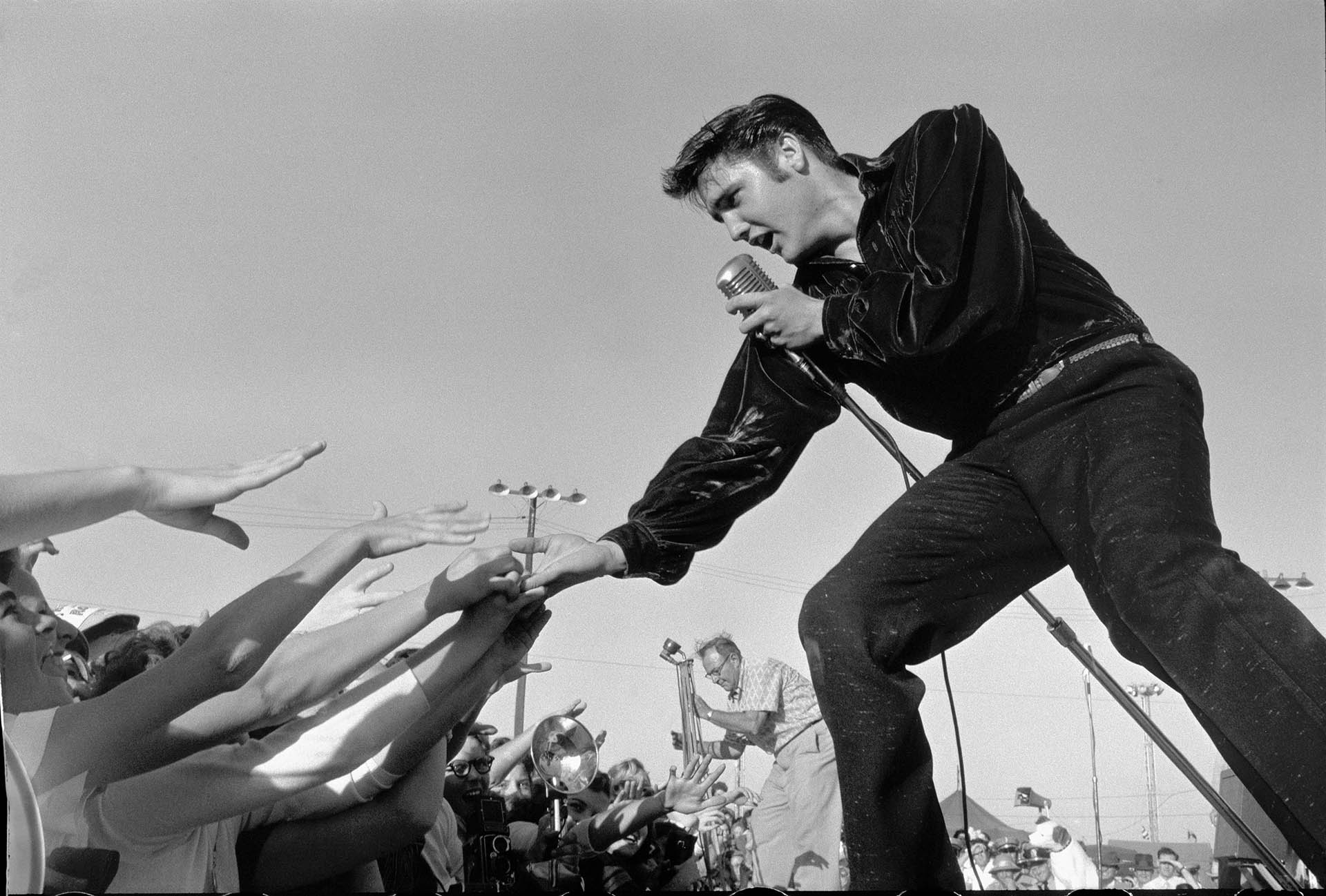 Elvis curvando-se para sua audiência em calças oxford e vestindo uma camisa de veludo, ele apareceu para dar um mega recital