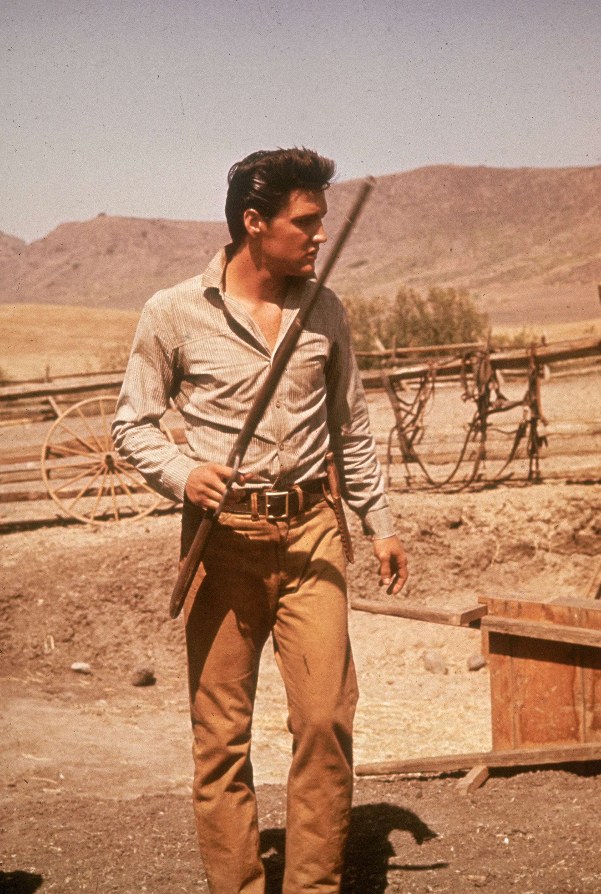 Além de ser um músico brilhante, ele se destacou em atuar. No set de filmagem completo, Elvis com uma espingarda na mão. Gabardine camel pants com cinto e camisa com decote em V (Foto de Hulton Archive / Getty Images)