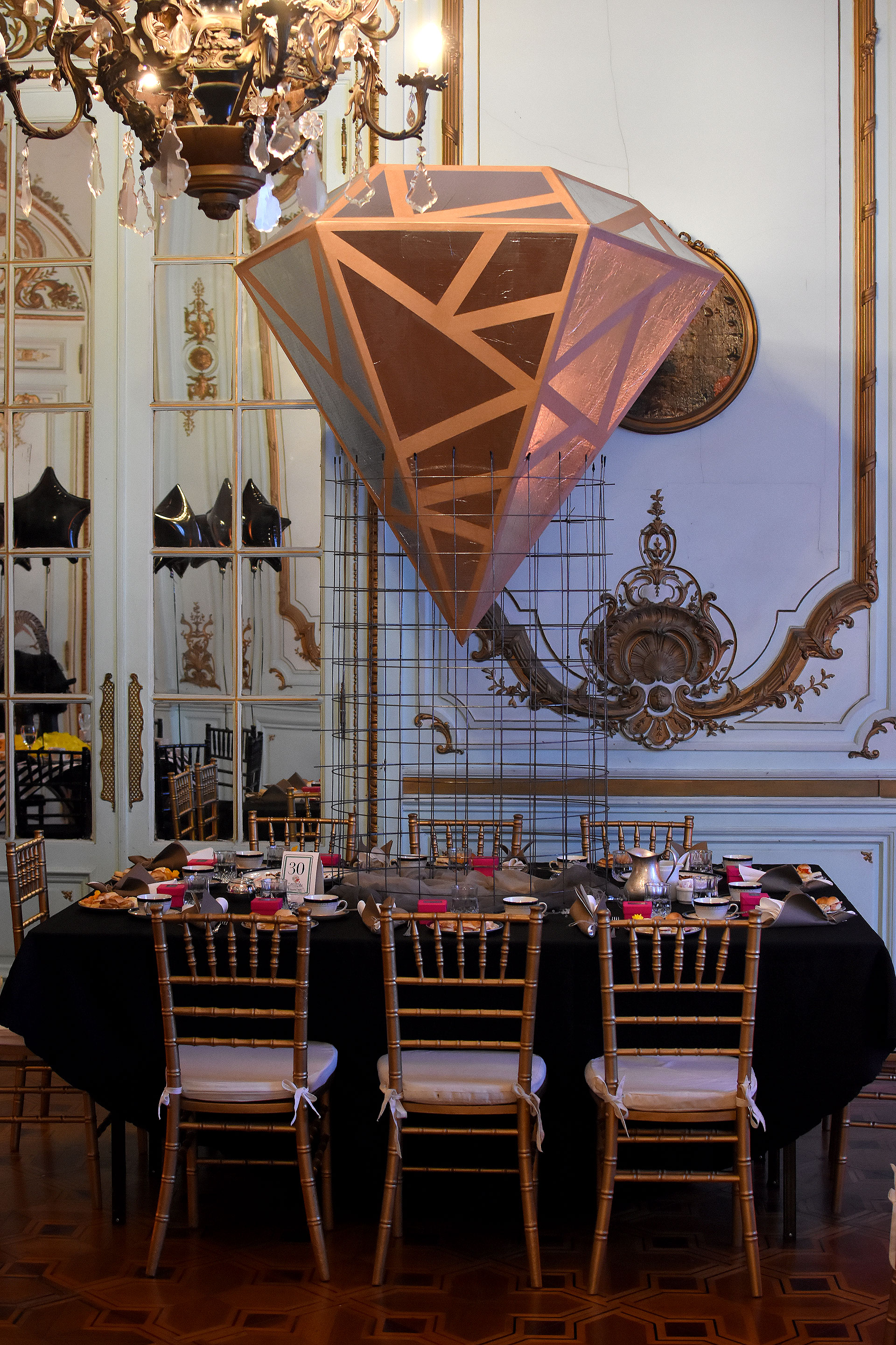“Alma D Diamante”. Una estructura de alambre sostiene al diamante dorado. Mantel y servilletas negras y grises. Detalles en fucsia y la elección de las sillas Tiffany