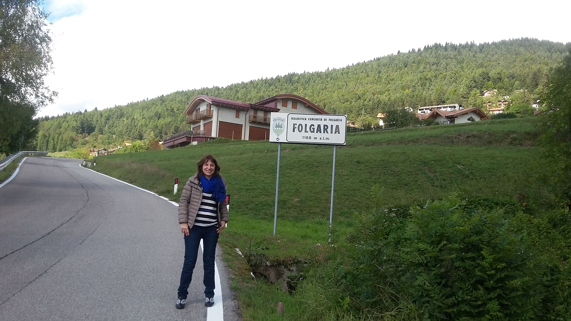 La autora de este texto, junto al cartel de la ruta que anuncia el nombre pueblo del norte de Italia.