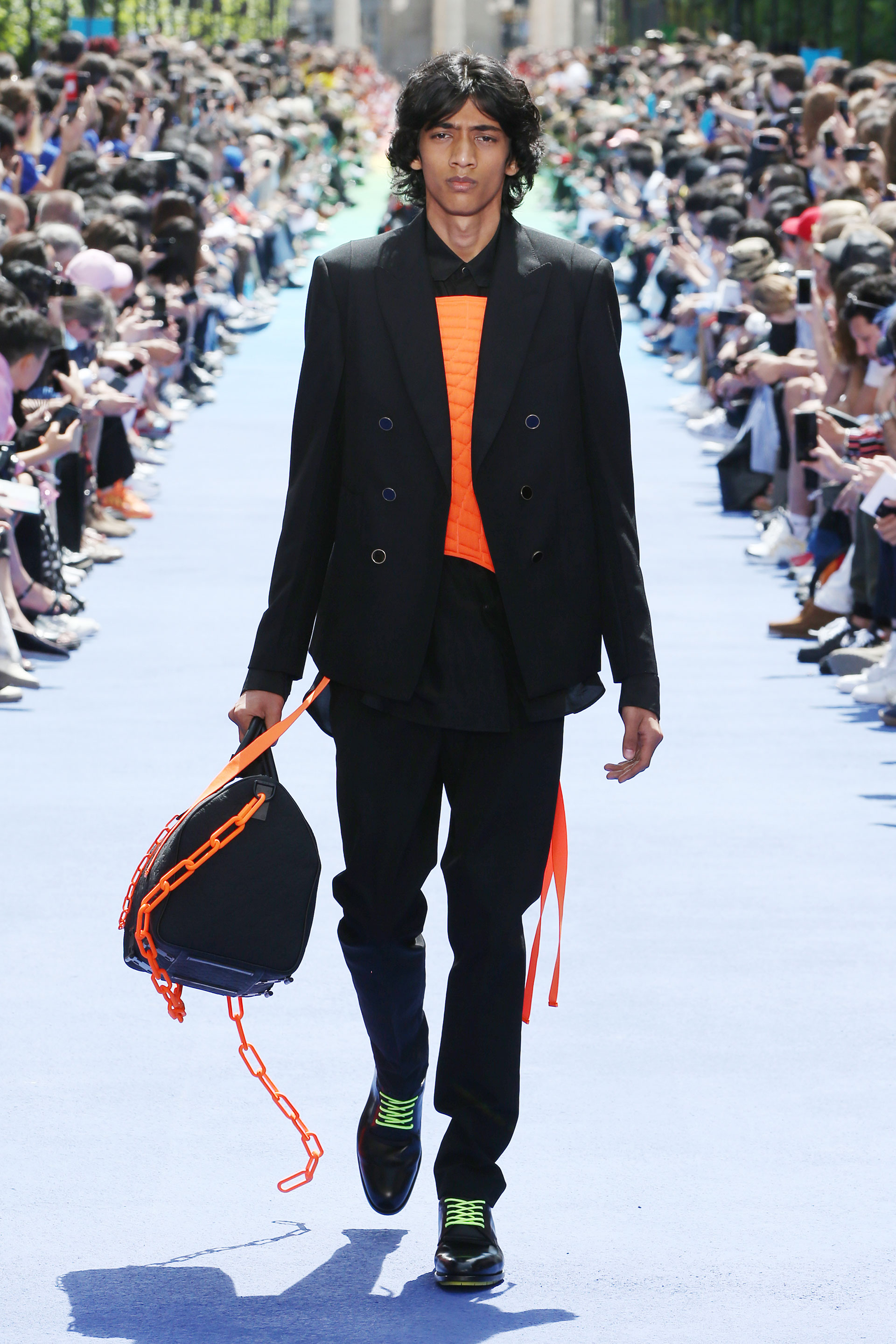 Naranja flúo y negro, una combinación de colores explosiva. Un pantalón de vestir chupín y blazer. Debajo, camisa negra con estampado naranja. Para acompañar el look, el bolso Keepall combinado con naranja flúo. ¿El detalle? Los zapatos negros con cordones verde flúo