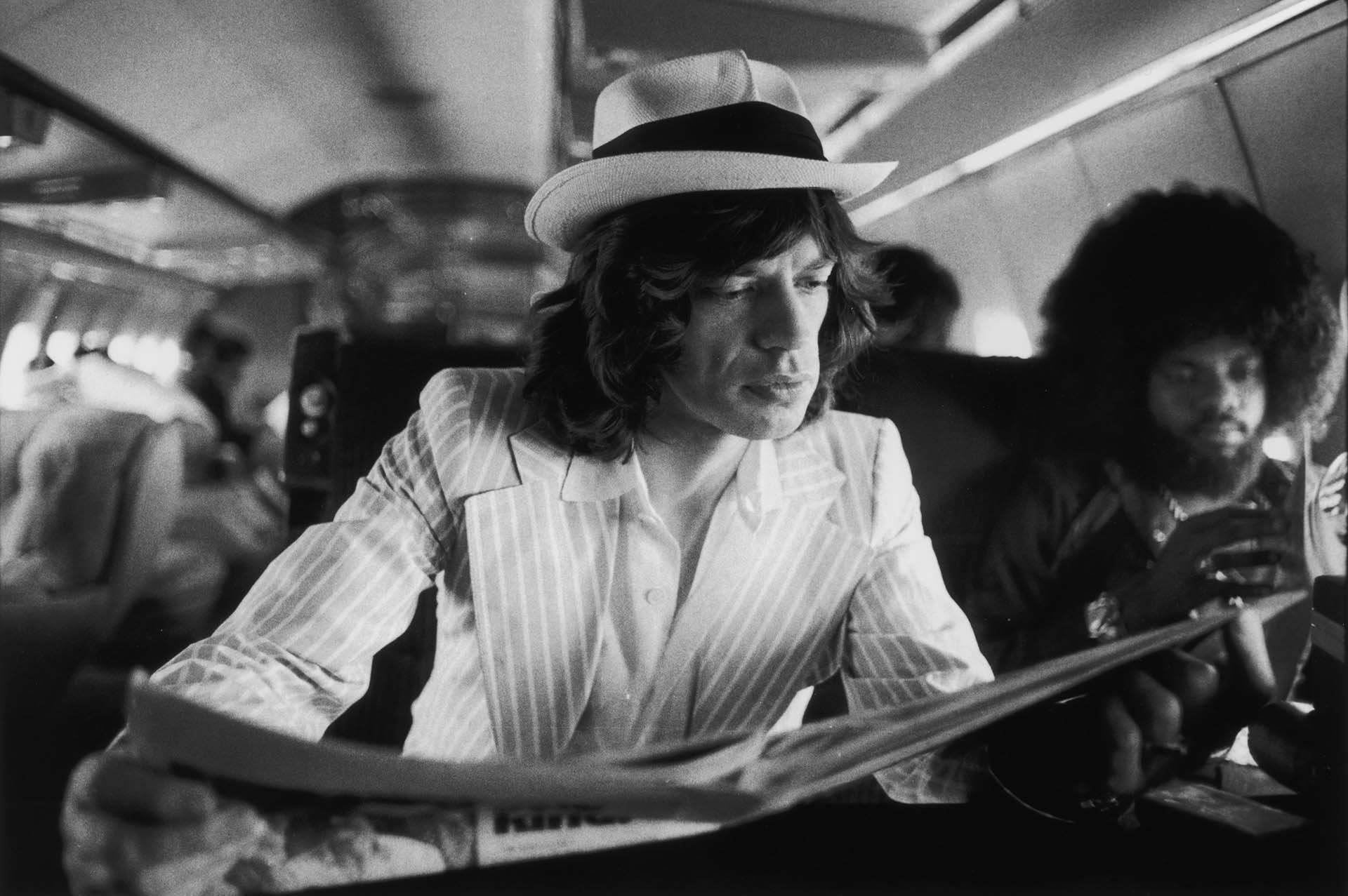 Mick Jagger lee los periódicos mientras viaja entre conciertos en el jet privado de los Rolling Stones durante su Tour of the Americas de 1975. También a bordo está el tecladista estadounidense Billy Preston (derecha).