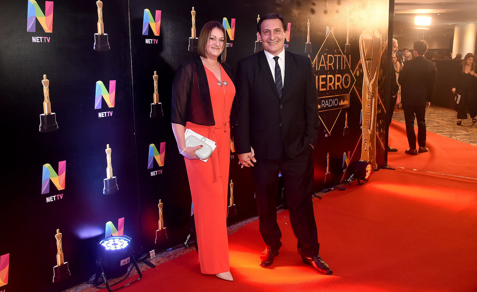Paulo Kablan con traje azul noche y zapatos en punta de cuero y su mujer Edith Garibotti con un mono colorado, solero de seda, clutch blanco y zapatos en punta nude