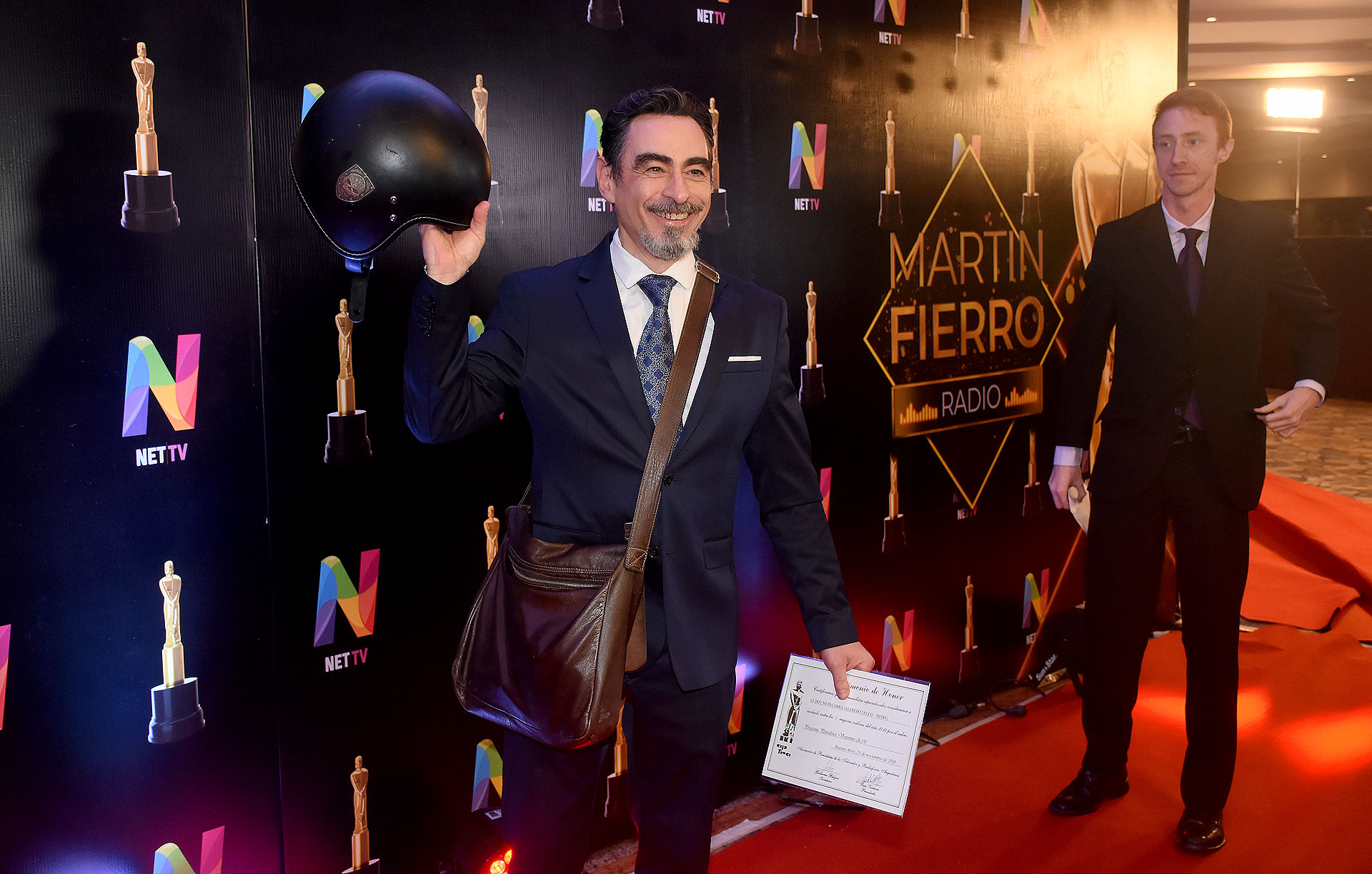 Federico Andahazi llegó a la alfombra roja con un traje azul, camisa blanca, corbata estampada y el detalle informal, el casco de la moto y un morral de cuero marrón