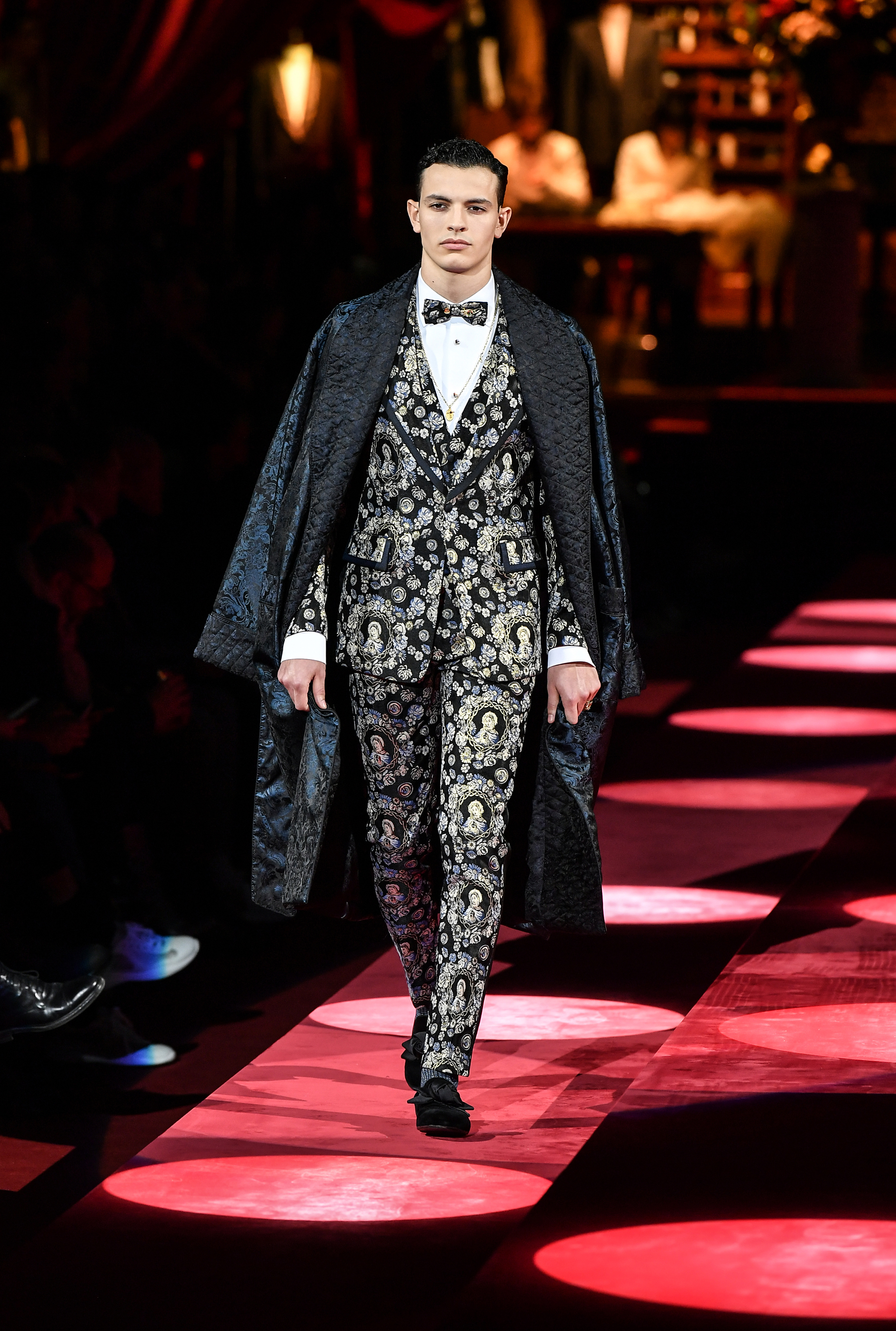 Uno de los looks más extravagantes del show. Con un sobretodo tornasol, y esmoquin con estampado de vírgenes y flores, al igual que el moño, con mocasines de raso con soquetes, camisa blanca con botones