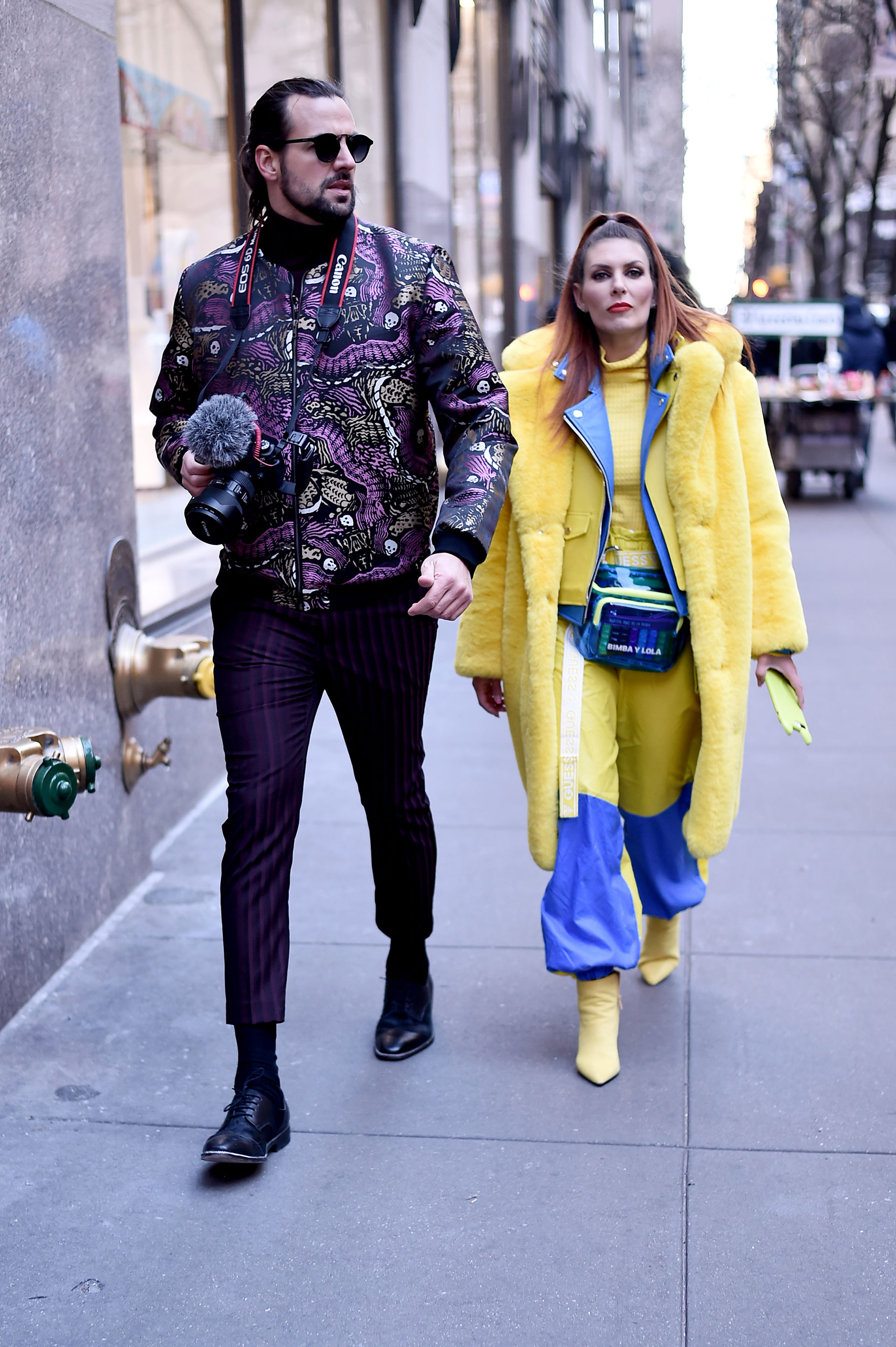 Dúo fashionista. Él con pantalón crop rayado de vestir en color púrpura, con zapatos acordonados y bomber estampada con animales. Ella con mix de texturas, piel, impermeable y cuero, todo en azul y amarillo. El detalle chic, la riñonera de pvc