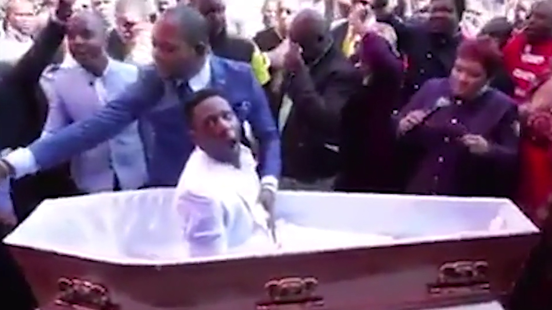 El pastor pone sus brazos sobre el pecho del hombre “muerto” y lo “resucita” durante la ceremonia en Sudáfrica, sin embargo el proveedor del funeral ha dicho que tomará acciones legales en su contra por dañar su imagen Foto: captura de pantalla.