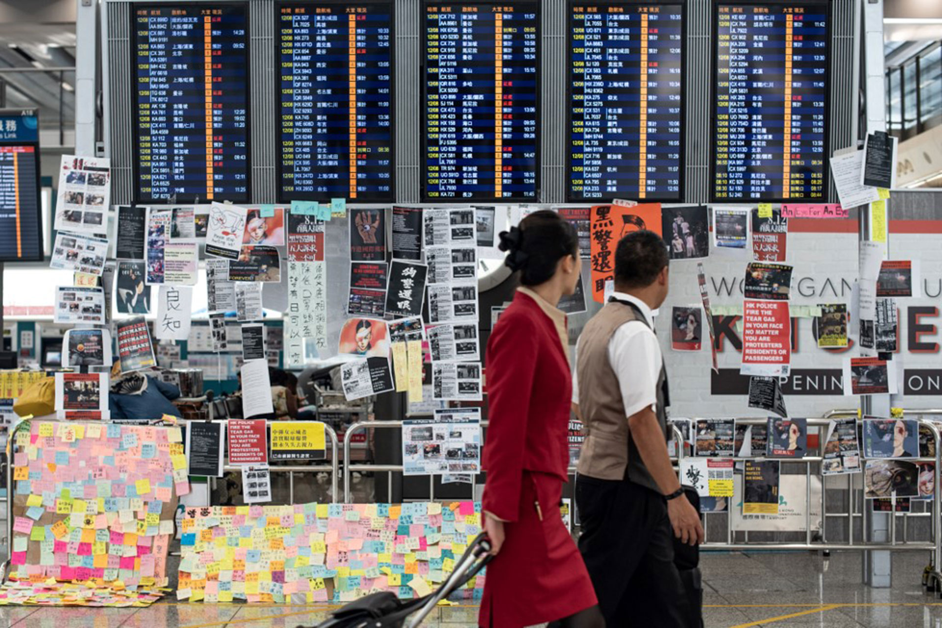 El 13 de agosto de 2019, al día siguiente de que el aeropuerto de Hong Kong cerrara sus puertas debido a las protestas a favor de la democracia, dos azafatas pasan por delante de un tablero de anuncios eléctrico cubierto de carteles (Foto de Philip FONG / AFP)