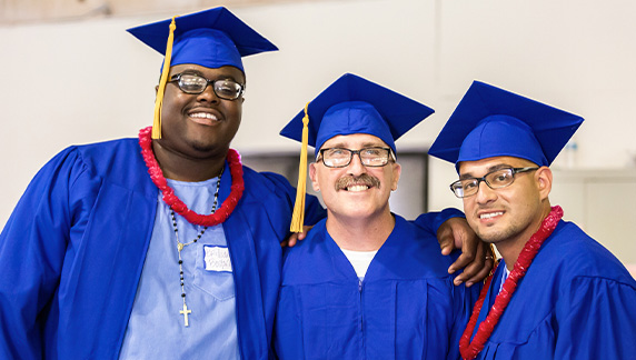 Three adult men smile proudly while wearing graduation robes.