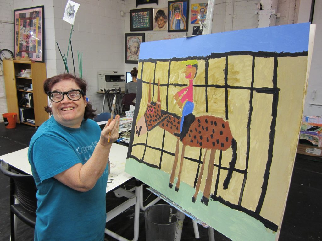 Woman holding a paintbrush in front of a large painting of a horseback rider and horse