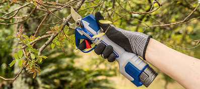 Mão calçando luva de proteção operando uma tesoura de poda a bateria Tramontina by TOTAL. A ferramenta, nas cores azul e cinza, está posicionada realizando o corte de um galho fino de árvore em um ambiente externo arborizado.