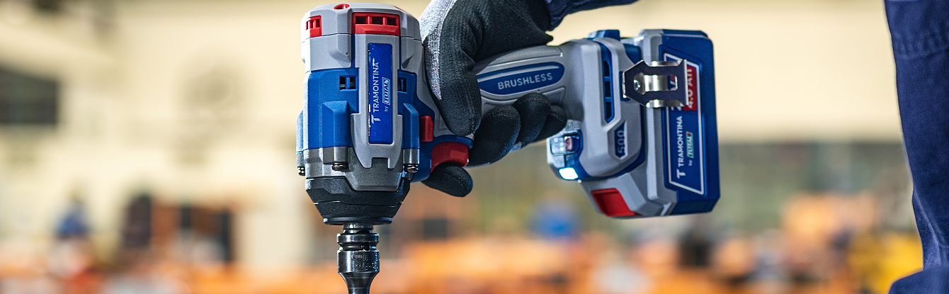 Banner horizontal mostrando em close uma mão com luva preta segurando uma chave de impacto Tramontina by TOTAL. A ferramenta exibe a inscrição "Brushless" no cabo, possui uma bateria de 4.0 Ah acoplada com indicador de carga LED aceso e um clipe de cinto 