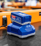Bateria Tramontina by TOTAL de 20V MAX e 4.0 Ah conectada a uma base de carregamento azul sobre uma bancada de oficina preta. A base possui indicadores de LED acesos e o cabo está conectado a uma tomada ao fundo em uma calha laranja.