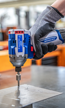 Close-up de uma mão calçando luva de proteção cinza e preta operando uma chave de impacto Tramontina by TOTAL. A ferramenta possui motor Brushless, design em azul e cinza, e está sendo utilizada para fixar um parafuso em uma placa de metal sobre uma banca