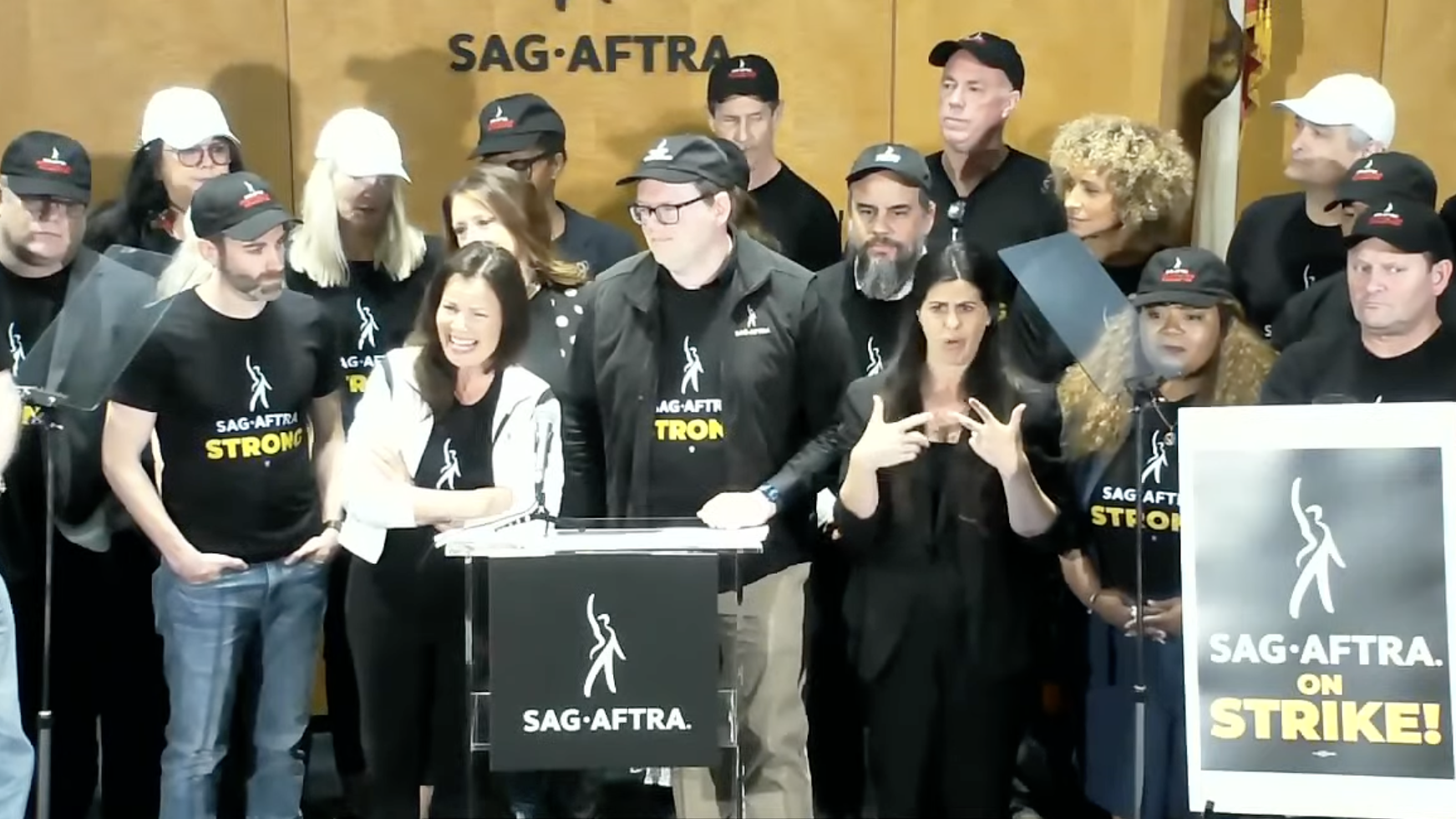 SAG-AFTRA National President Fran Drescher, centered right, and Chair of Negotiating Committee Duncan Crabtree-Ireland, standing to her left speak at the SAG-AFTRA press conference announcing an official strike on Thursday, July13, 2023 in Los Angeles. Screenshot: YouTube/SAG-AFTRA