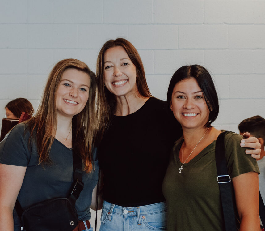 Three young ladies smiling and standing together with their arms over each other's shoulders.