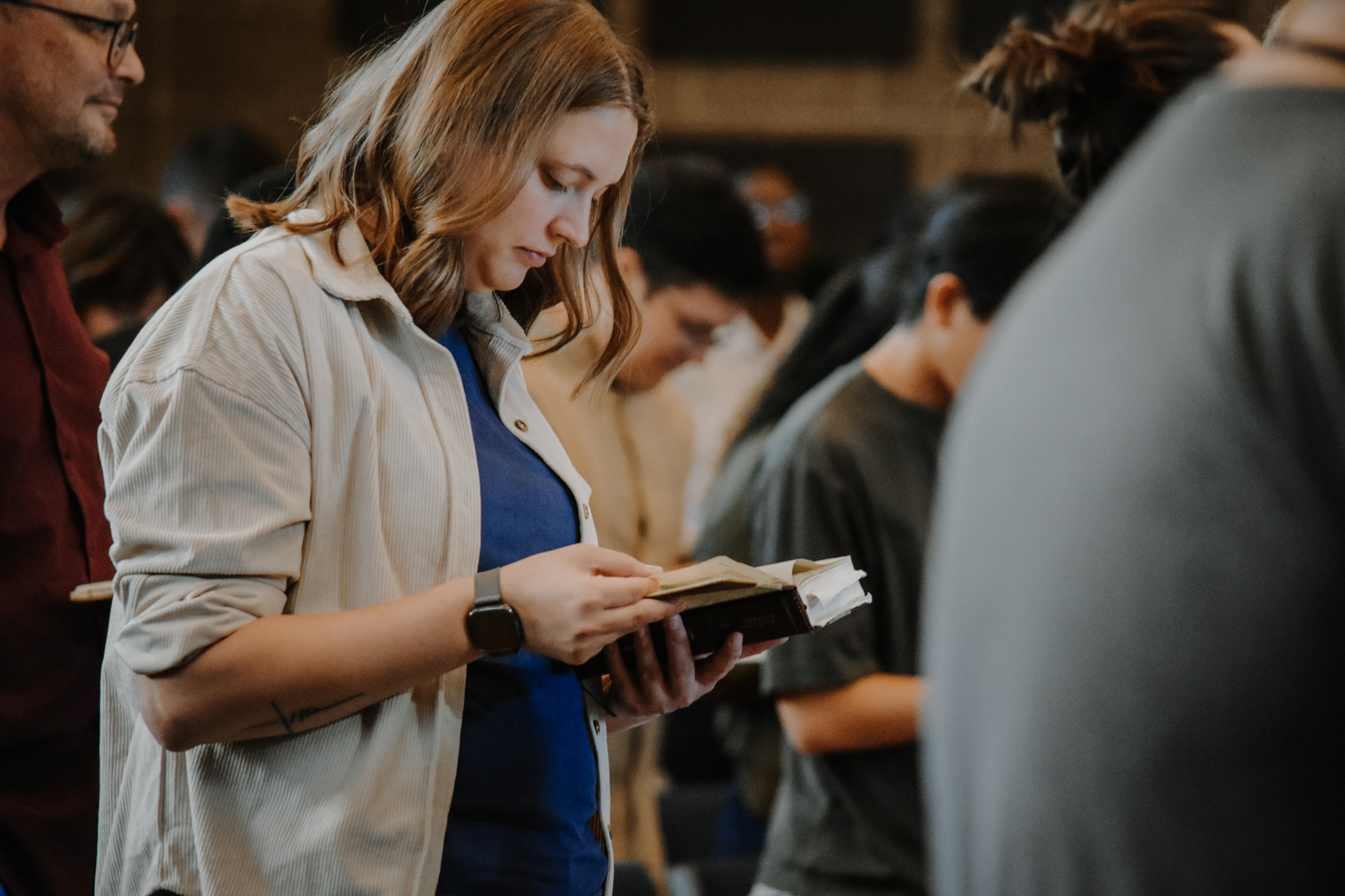 A young lady is standing amongst a congregation of people, holding their Bibles open and reading them.