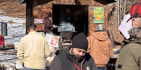 A man holding a log in front of a maple shack on a chilly day.