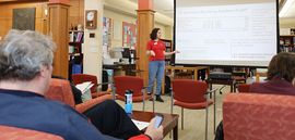 A person standing in front of a presentation screen, speaking with a small audience.
