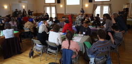 A large indoor space with people sitting around tables listening to a speaker.