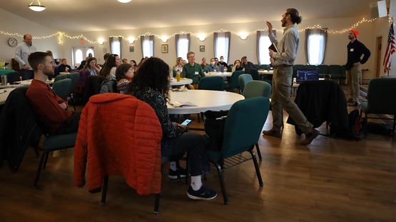 Two people holding a presentation for onlookers sitting at round tables inside a community center. 