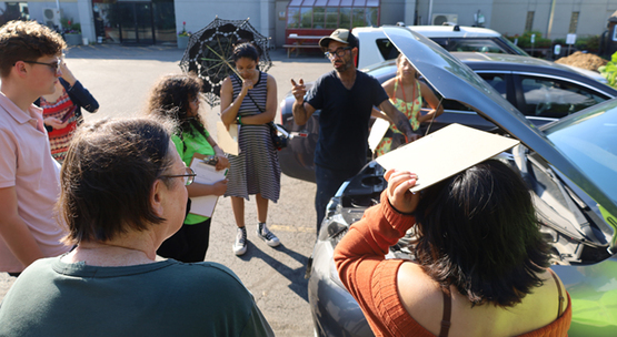 In a parking lot, a group of people gathered around a car as a mechanic explains what's under the hood.