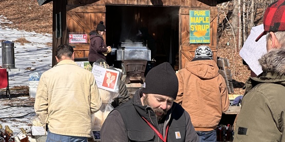 A man holding a log in front of a maple shack on a chilly day.
