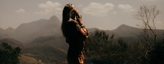 A woman standing outside looking to the sky with her eyes closed and her hands to her chin with mountains and trees behind her. 