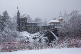 Belvedere castle surrounded by rocks and trees covered with snow, and a small body of water in front of it.