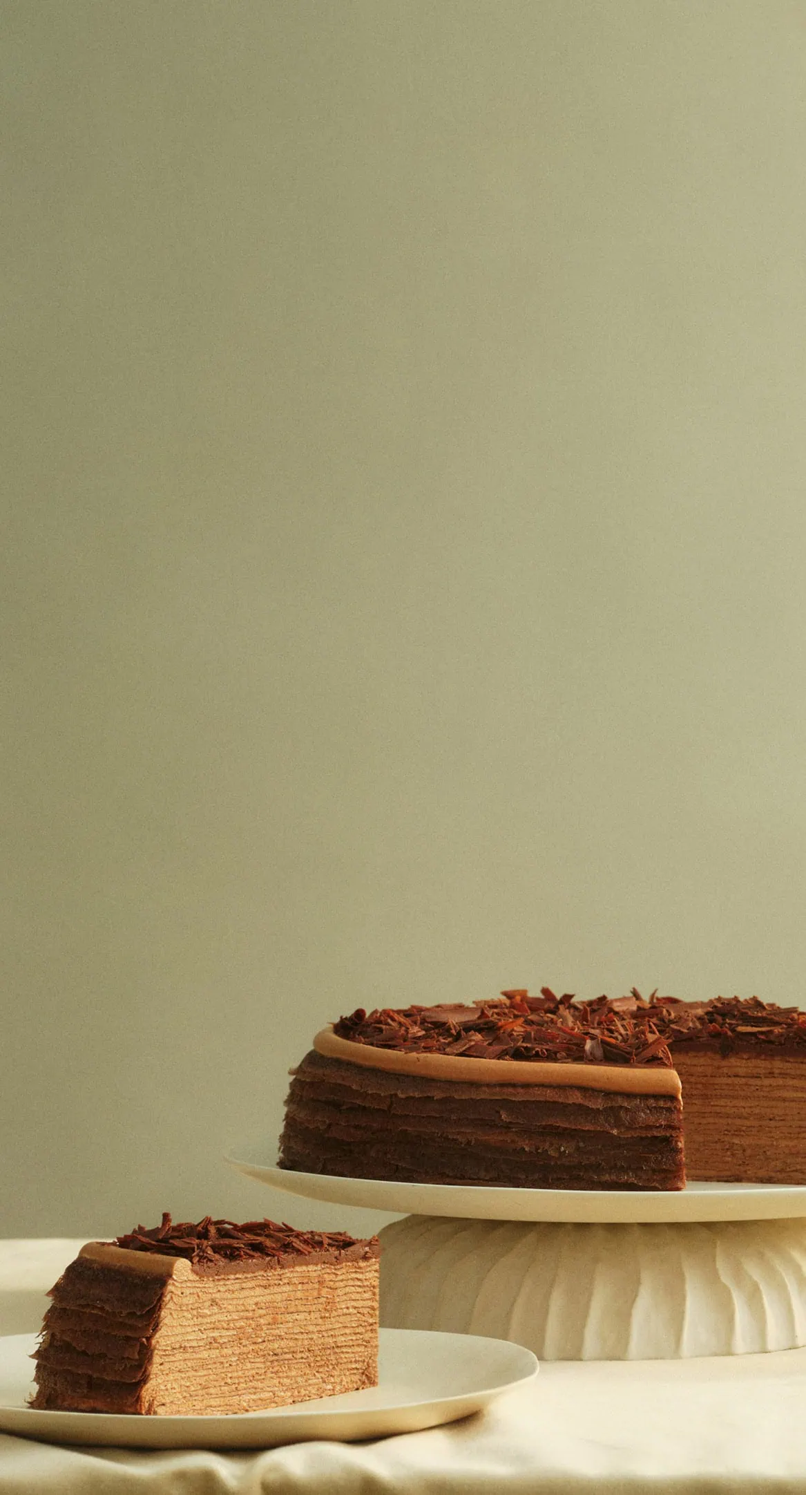 A slice of Amedei Chocolate Mille Crêpes sits on a white plate next to the full cake, displayed on a cake stand, against a soft beige background.