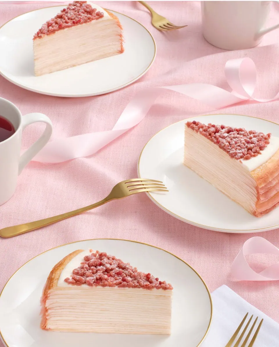 Three slices of Pink Pearl Mille Crêpes on a pink tablecloth, accompanied by forks and cups.