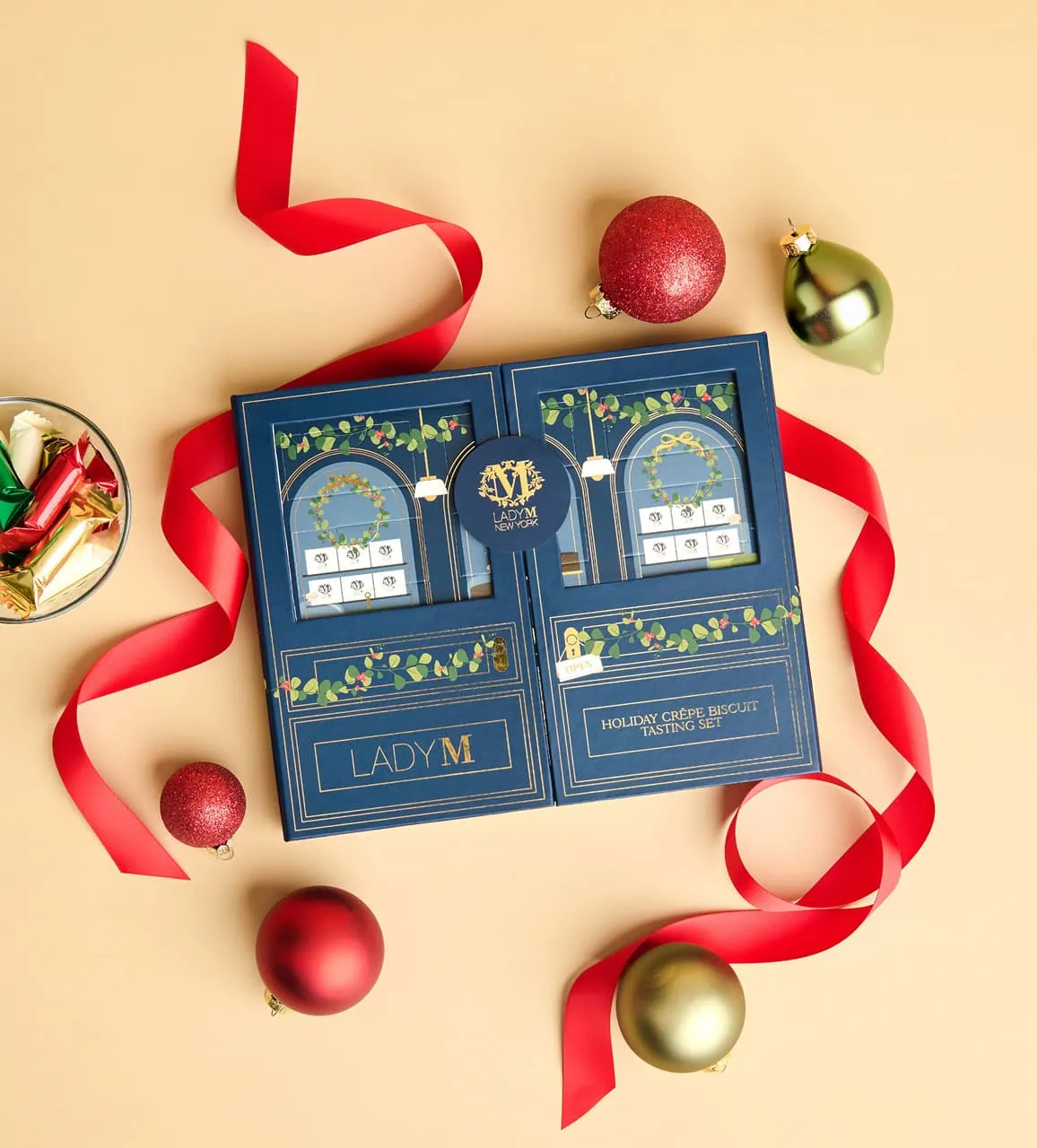 Lady M Holiday gift box surrounded by festive red and gold ornaments with red ribbon, ready for gifting