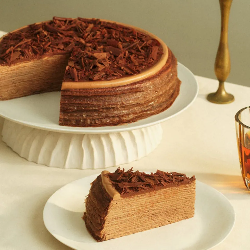A layered chocolate cake topped with chocolate shavings, with a slice on a separate plate beside it.