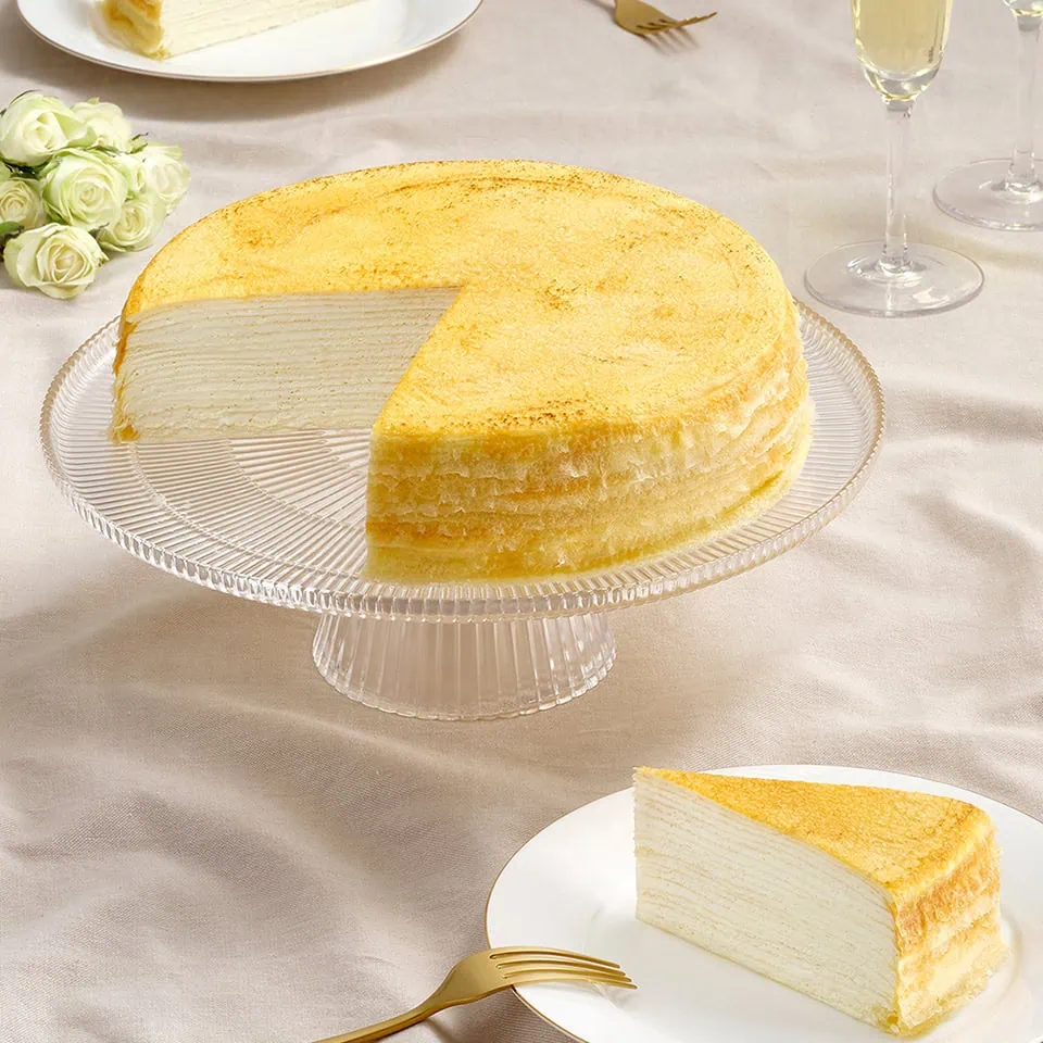 A layered cake on a glass stand, with a slice removed and placed on a plate. The setting includes a fork, a glass of champagne, and white roses.
