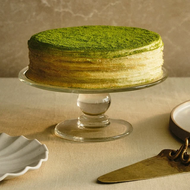 A matcha cake on a glass cake stand, with a green powdered top, placed on a linen surface alongside a knife and a small dish.