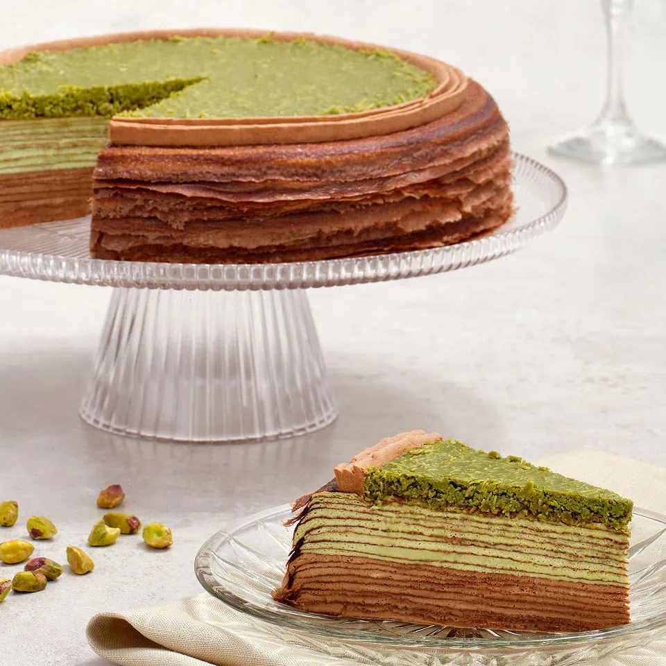 Two round cakes on display: one with a green matcha topping and the other a chocolate mille crêpe cake. Slices are plated alongside with pistachios.