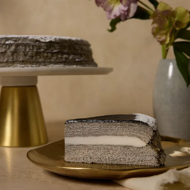 Sesame Mochi Mille Crêpes on a gold cake stand, with a slice on a gold plate showing thin gray crêpe layers, a mochi center, and a black sesame glaze