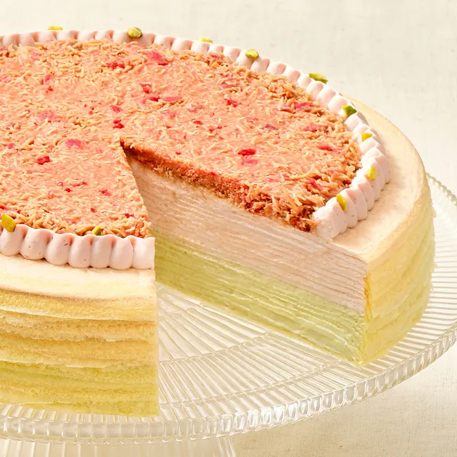 Dubai Strawberry Mille Crêpes on a glass cake stand with a slice cut away, showing pink strawberry and green pistachio layers under a crispy kataifi topping, edged with piped strawberry cream and chopped pistachios