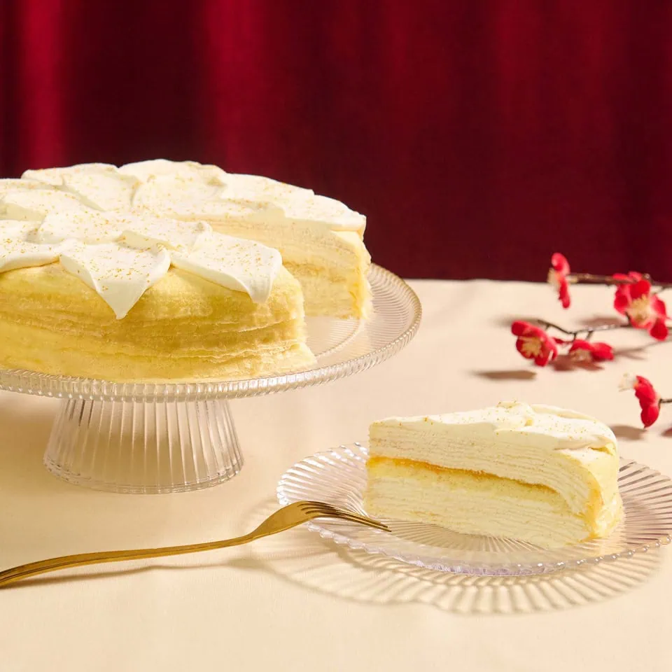A slice of layered cake topped with cream sits on a plate beside a whole cake on a glass stand, with decorative flowers and a gold fork.