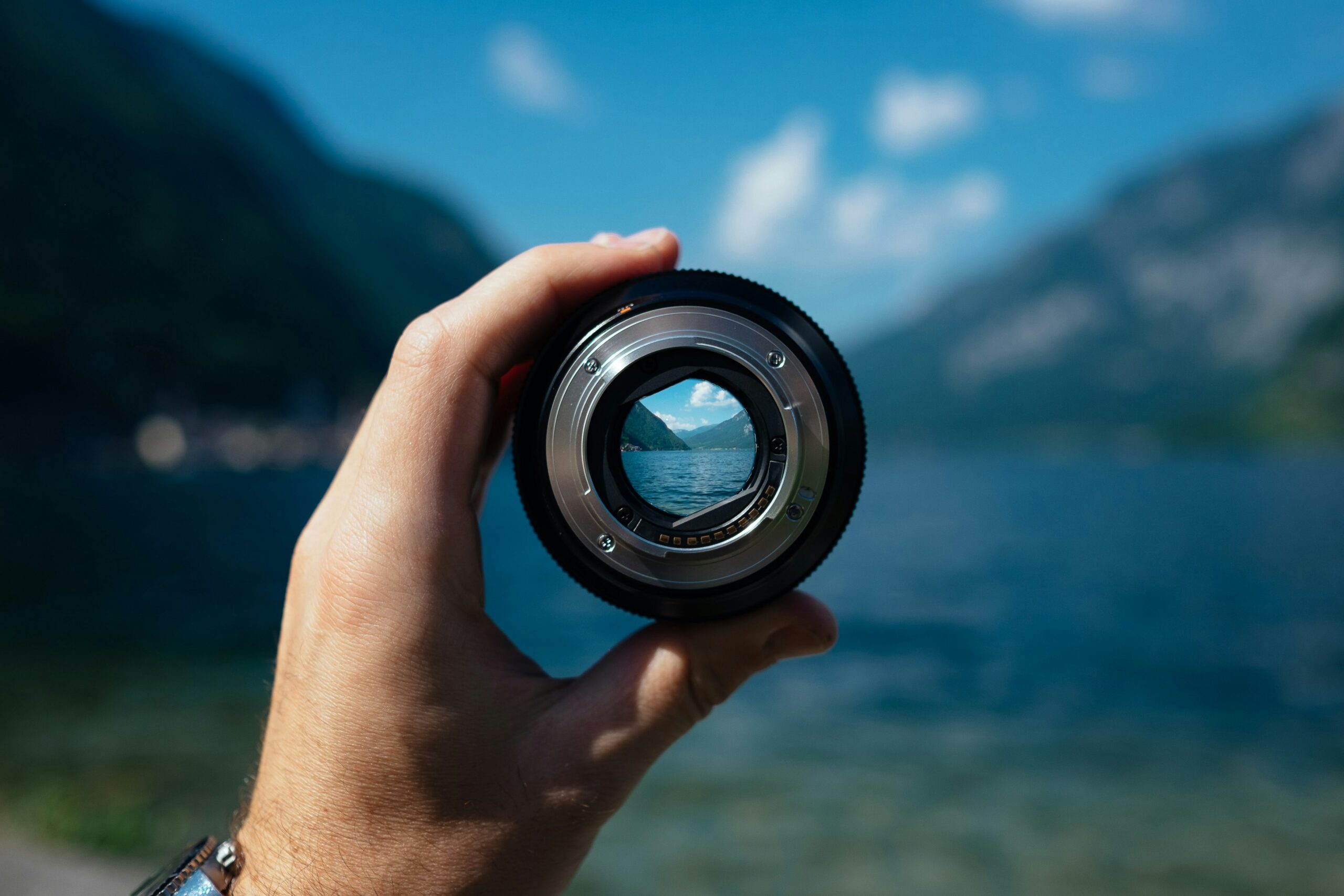 a hand holding up a magnifier or scope in front of water and mountains behind