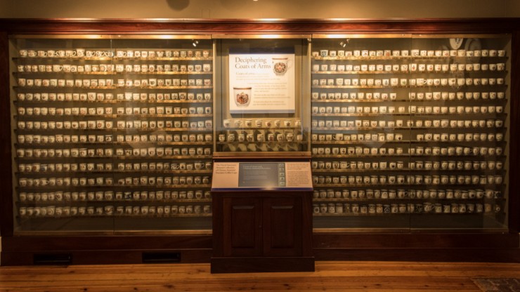 A wall of tea cups, all with faint coats of arms on them, as part of an exhibit titled "Decipering Coats of Arms"