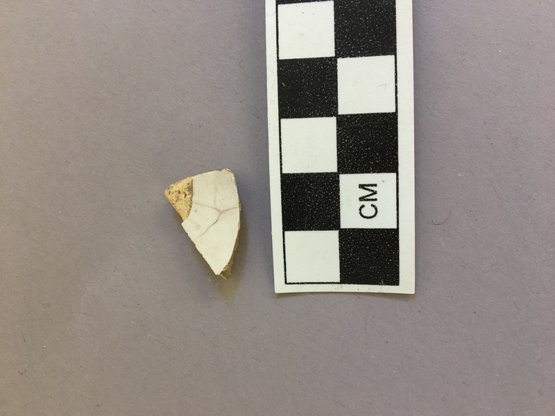 Tin-glazed earthenware sherd. This ceramic sherd is distinguishable as tin-glazed earthenware by its buff pasty and shiny, fragile glaze. It is 1.9 cm long, 1.2 cm wide, and 0.4 cm thick.
