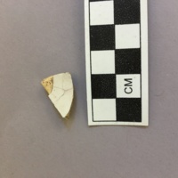 Tin-glazed earthenware sherd. This ceramic sherd is distinguishable as tin-glazed earthenware by its buff pasty and shiny, fragile glaze. It is 1.9 cm long, 1.2 cm wide, and 0.4 cm thick.