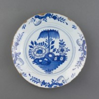 English delftware plate from the turn of the 17th-18th century, made from red earthenware with a tin-glaze overlaid, and a blue floral leaf border surrounding flowers under an umbrella-like tree.